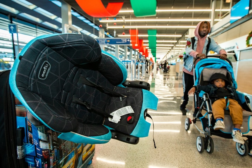 A car seat sits atop a pile of luggage as a man passes by, pushing a child in a pram.