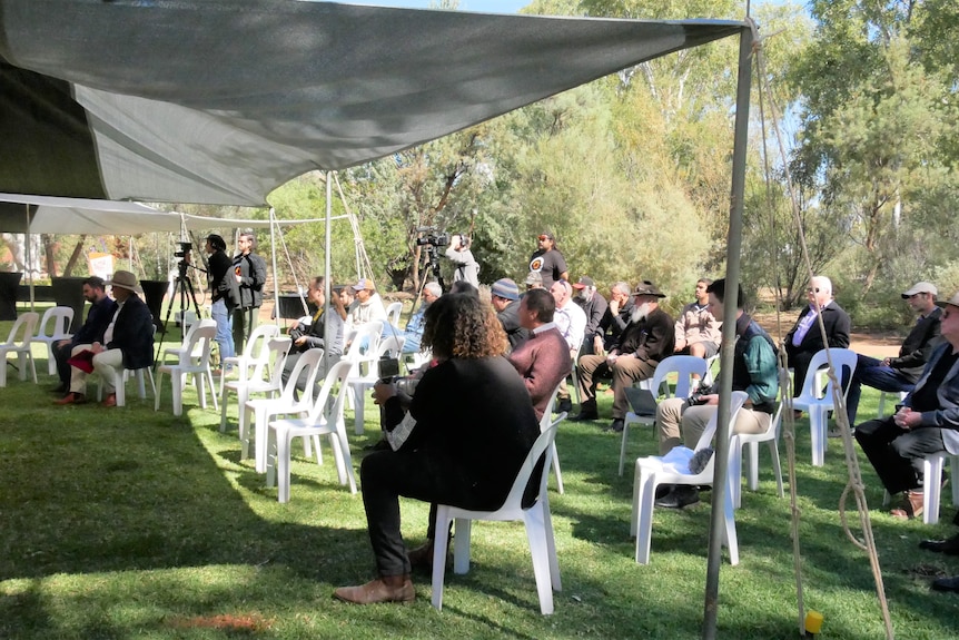 A group of people sit on plastic chairs on a grass area.