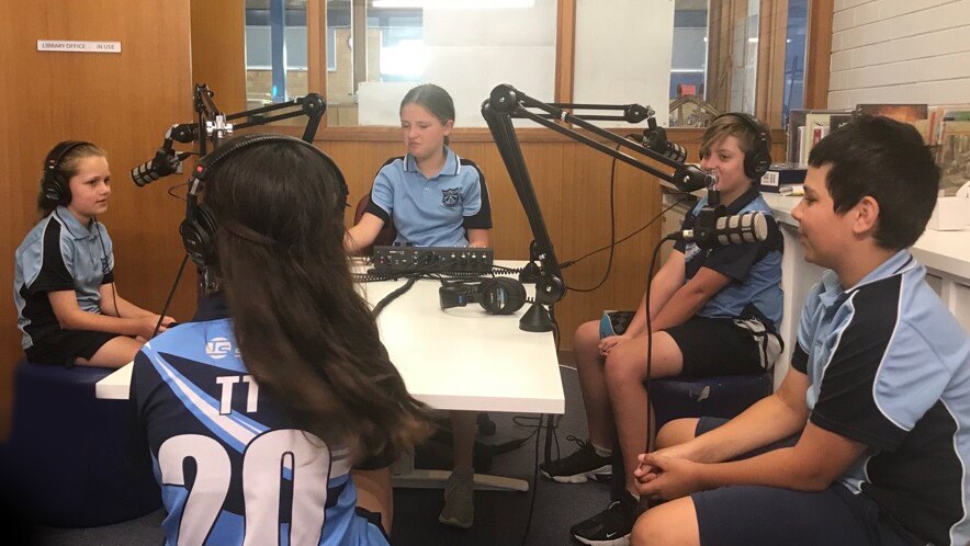 Microphones and recording equipment on a table and five students making a podcast.