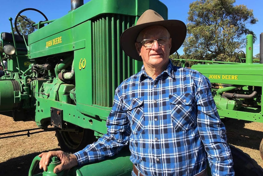 A man in a hat standing in front of two large green vintage John Deere tractors