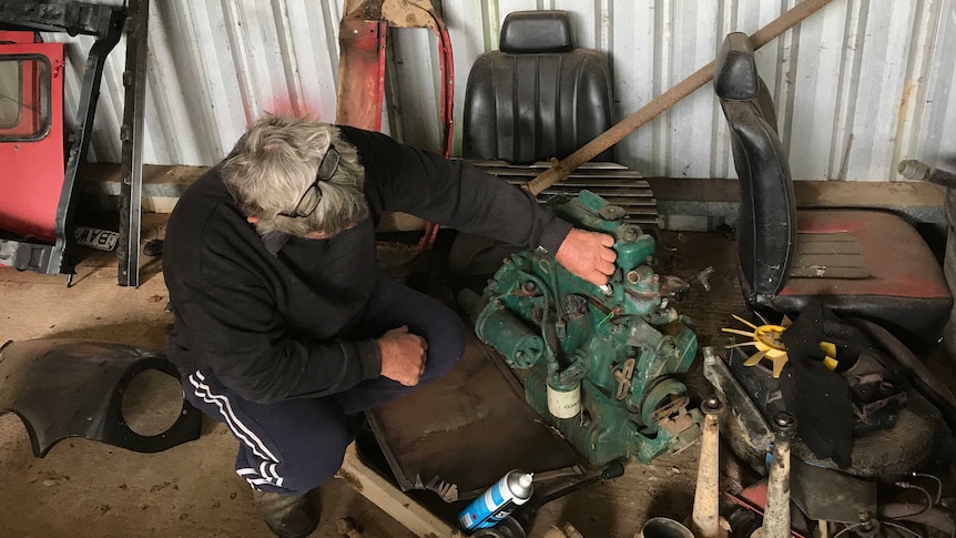 A man squats down in his shed, and fixes an engine on the floor