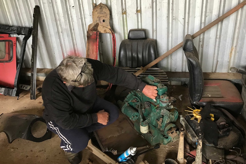 A man squats down in his shed, and fixes an engine on the floor