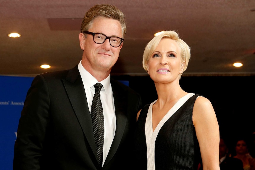 Left: Joe wears black suit and tie, black-rimmed glasses. Right: Mika wears black-and-white dress, has short blonde hair