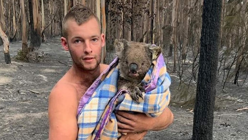 Patrick Boyle hugs a koala swaddled in a blanket, standing amongst burnt trees.