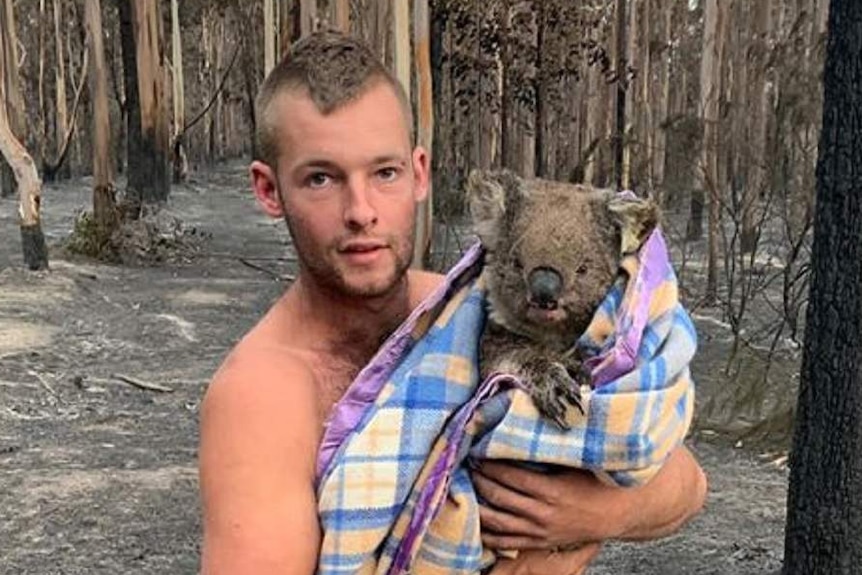 Patrick Boyle hugs a koala swaddled in a blanket, standing amongst burnt trees.