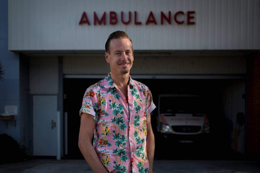 A man dressed in a pink shirt with a palm tree and flower print stands outside an ambulance station.