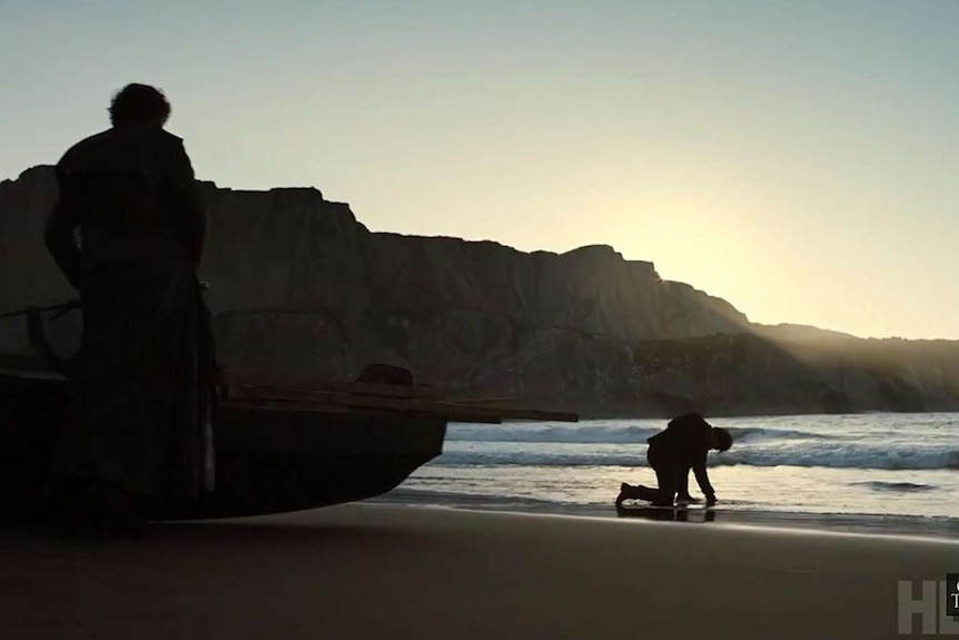 Theon Greyjoy bent over in the sand of a beach, recovering from a fight.