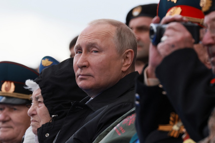 Vladimir Putin stares at the camera among a crowd of people watching a parade.