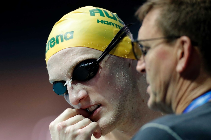 Mack Horton chews on his finger as water drips off his face while his coach looks on