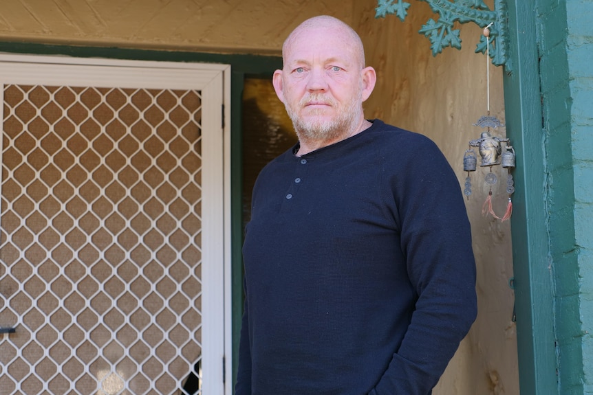 A man with a neutral expression on his face wearing a black jumper stands on the front porch of a home, looking at the camera.