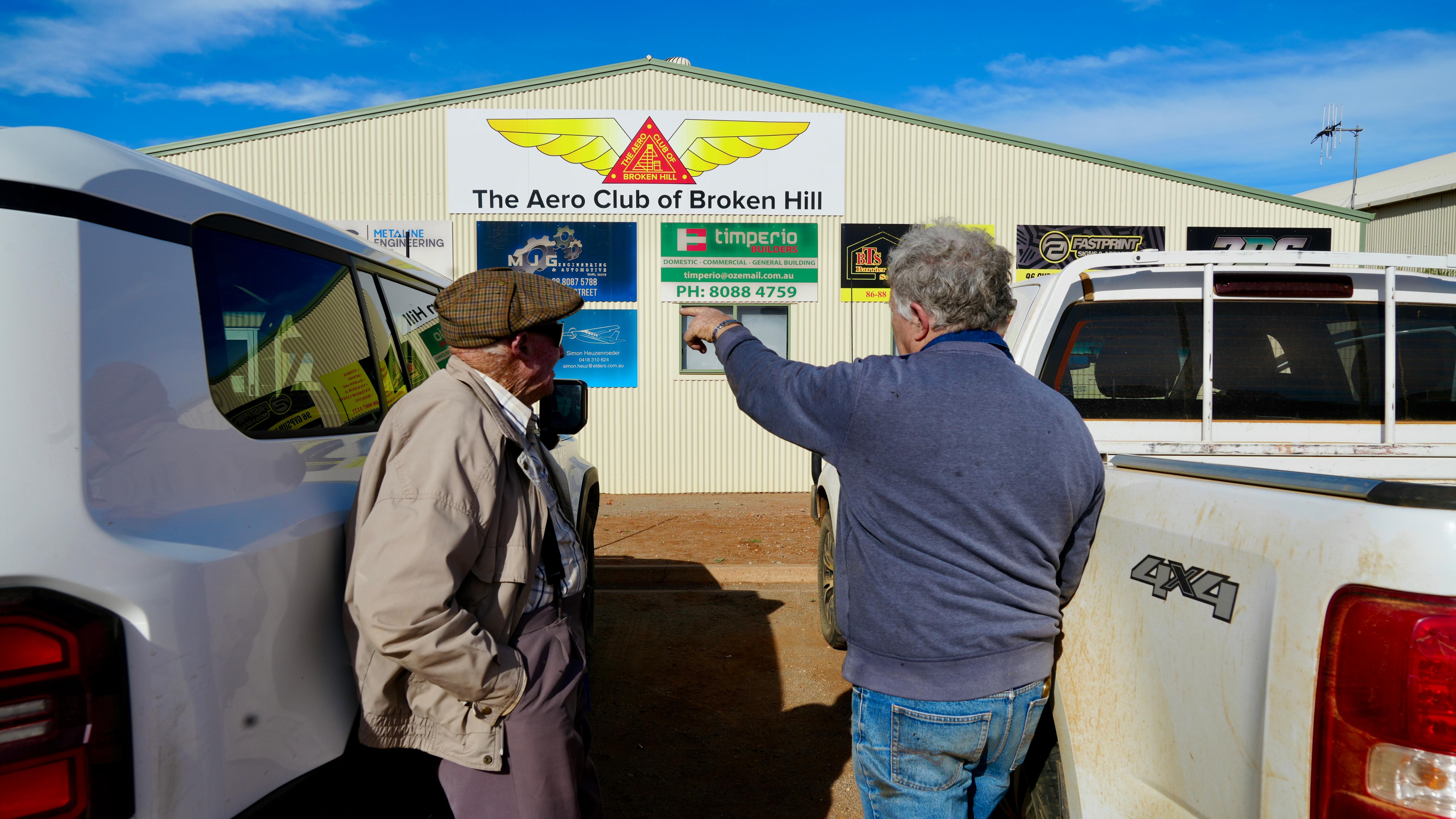 a man with a hat and another man with a blue jumper pointing to a shed with signs on it 