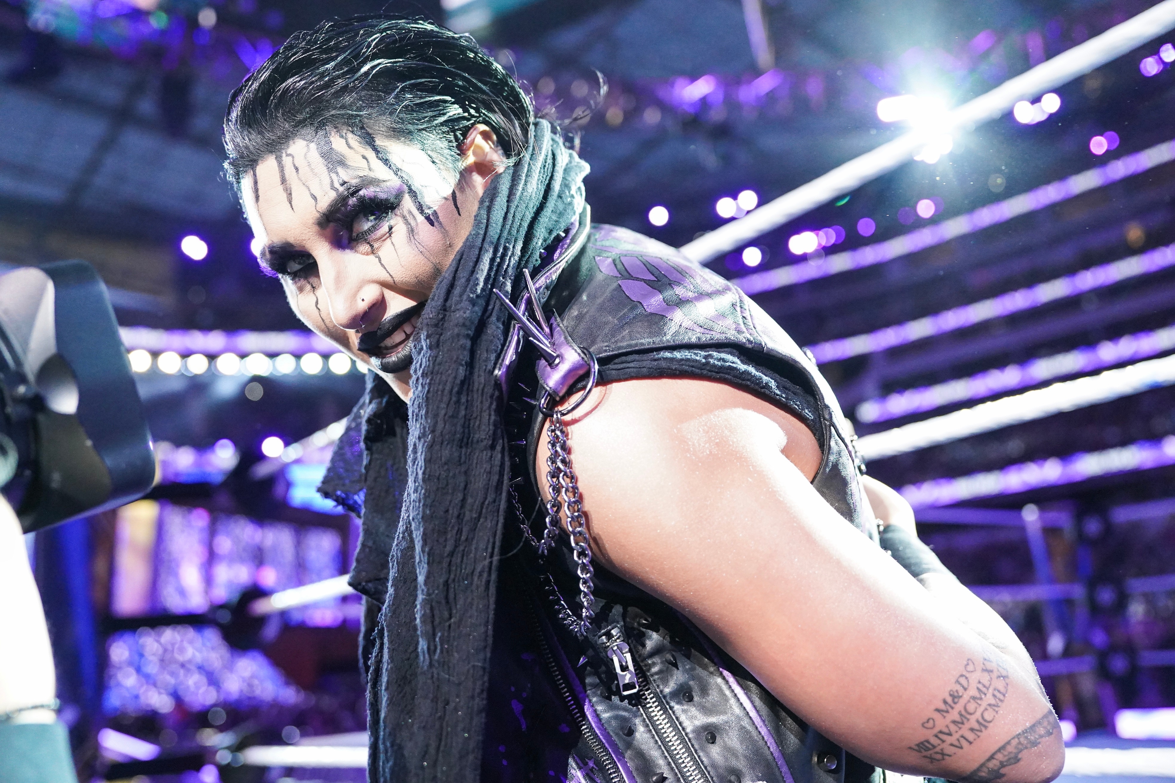 A woman wearing black clothing with purple shoulder spikes crouches on the outside of the ring mat glaring at the audience