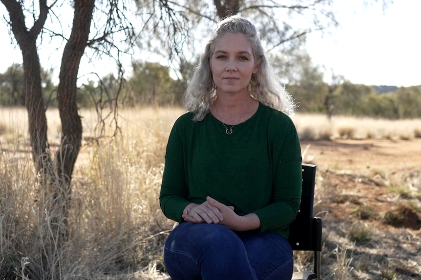 A woman sits on a chair outdoors, behind her is a tree and a dry grass clearing. She has a neutral expression.