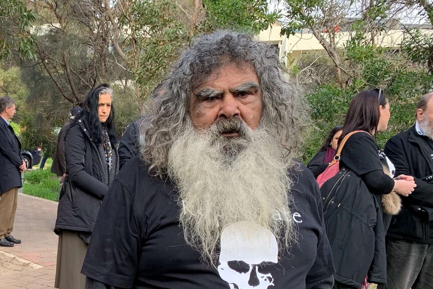A man with a big long white beard stares at the camera, he's wearing a t shirt with a skull on it saying Our Ancestors