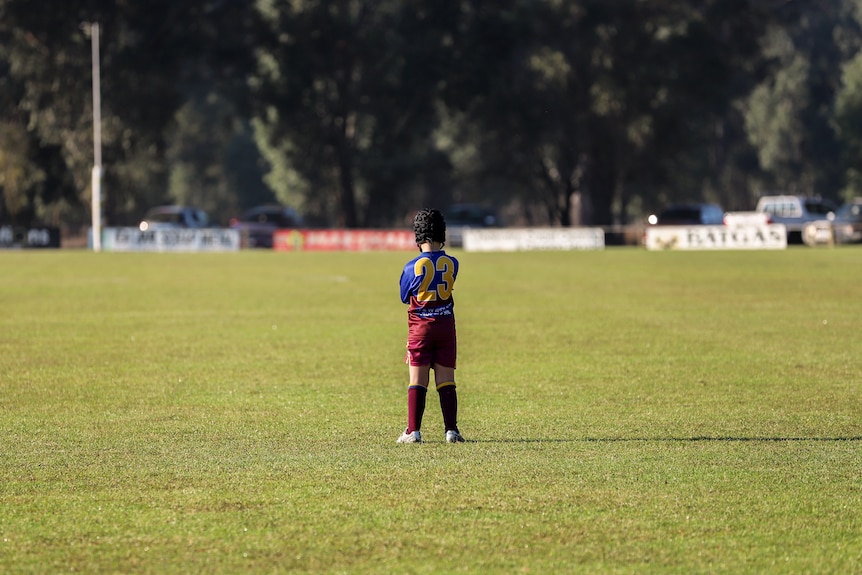 Young boy wearing helmet and football uniform stands on green grassed football oval