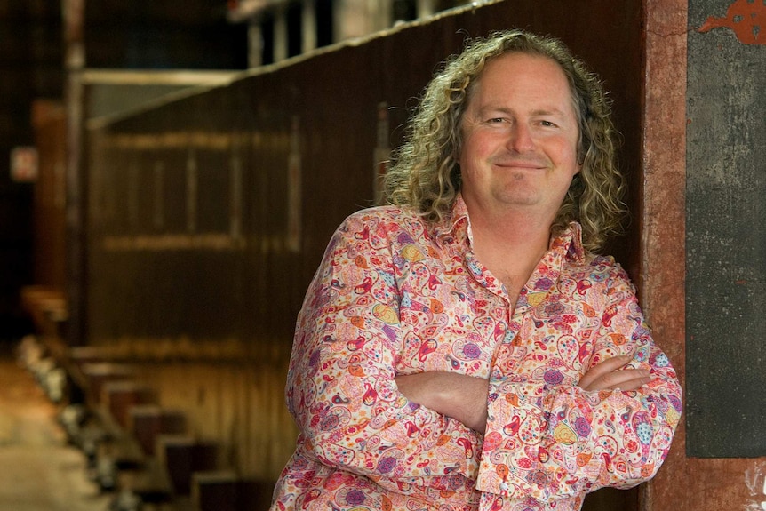 A man in a colourful shirt leans against a wooden wall and smiles at the camera.