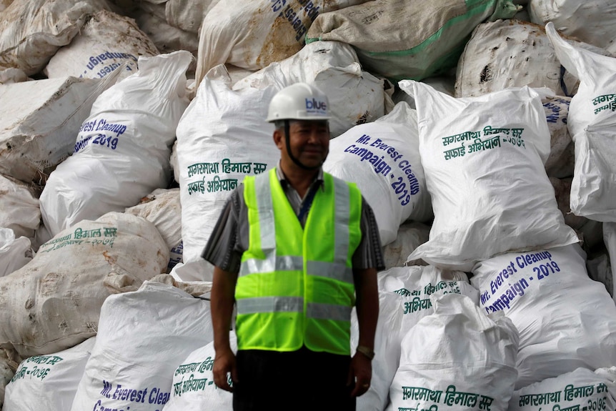 A man stands in front of a pile of garbage in white bags from Mount Everest.