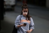 A woman wearing a mask looks down at her phone while walking.