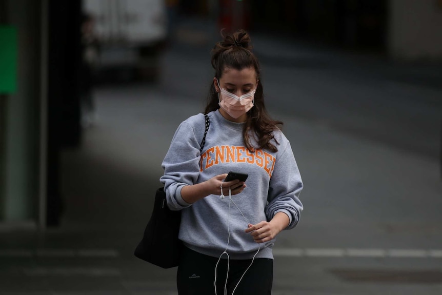 A woman with her brown hair half-tied up wearing a mask looks down at her phone with earphones dangling.