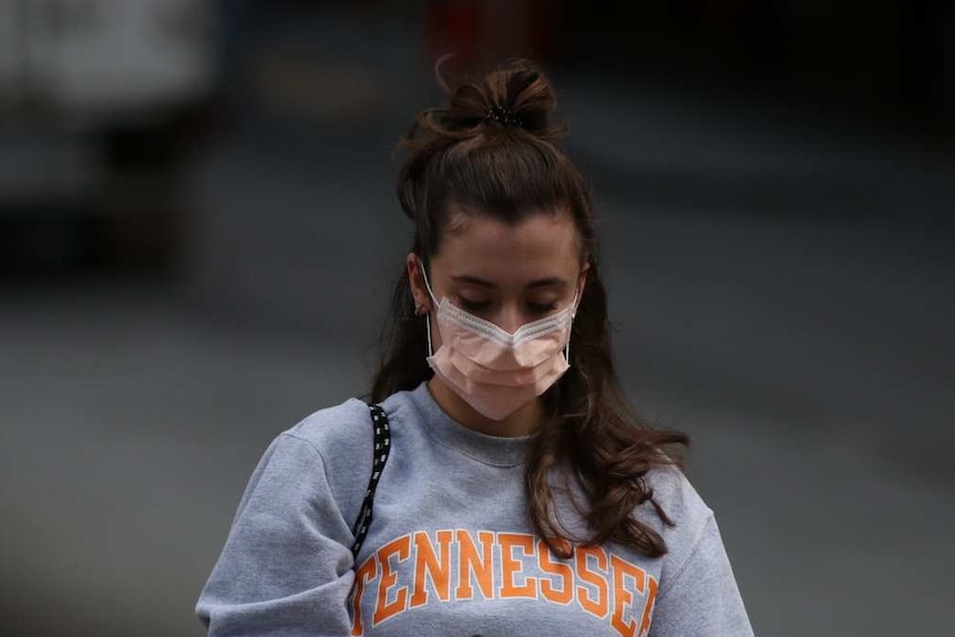 A woman wearing a mask looks down at her phone while walking.