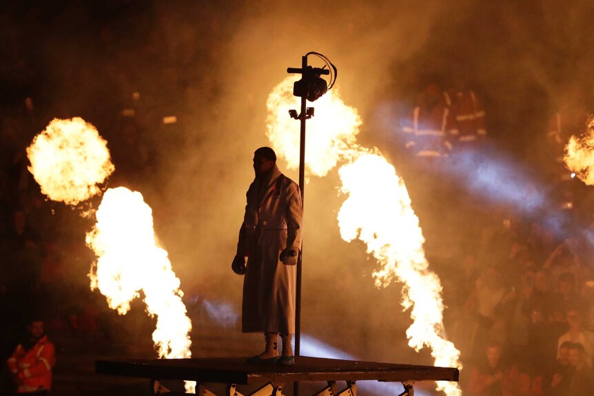 Anthony Joshua stands silhouetted b two pillars of flame while wearing a dressing gown and boxing gloves.