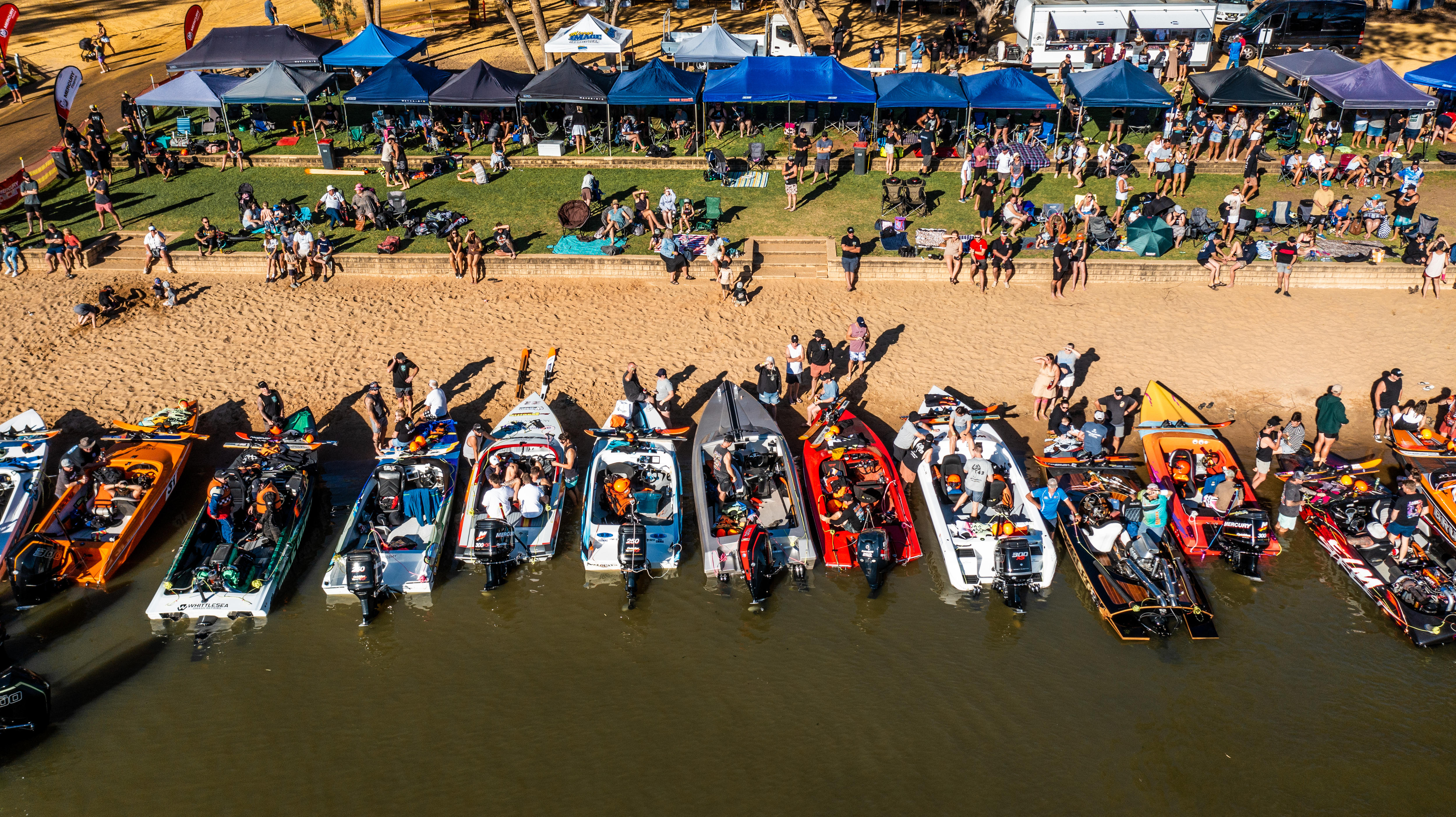 13 barcos de esquí acuático apuntando hacia un banco de arena con gente sentada en el césped cercano y bajo pabellones azules