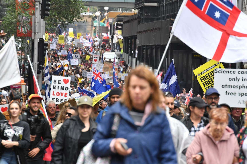 a crowd of people, some holding signs and flags, march through a street in Sydney, New South Wales