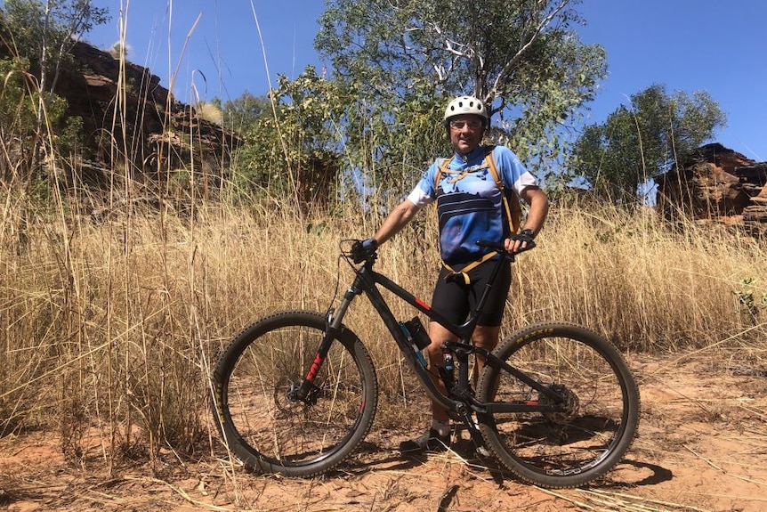 Man stands holding mountain bike and wearing helmet in front of tall dry grass