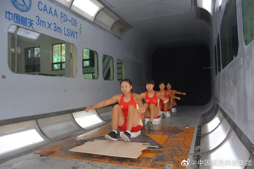 Four female rowers in red kit mime the actions of rowing while sitting in formation in a wind tunnel.