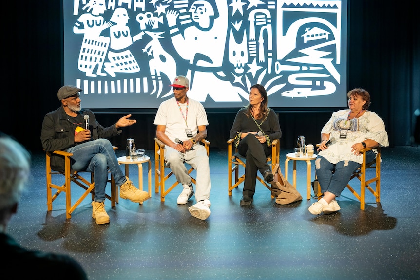 Four people in discussion sitting on chairs on stage, the Birrarangga artwork behind them