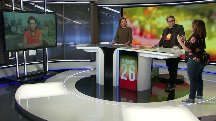 Two women and a man standing at a desk in a TV studio with another woman on a TV screen.