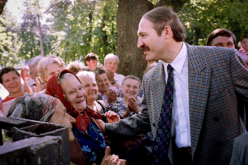 A moustachioed man in a suit greets older women with scarves tied around their heads