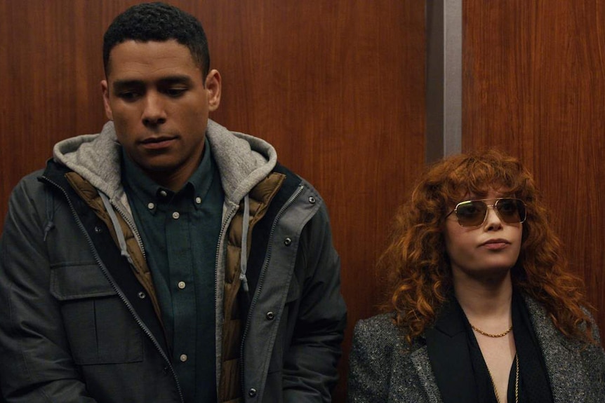 man and woman stand beside each other in wood-panelled elevator, looking awkward.