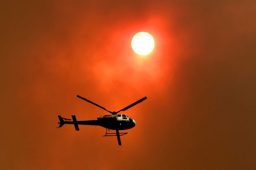 A helicopter flies through a dark, reddish sky with the sun partially obscured by smoke.