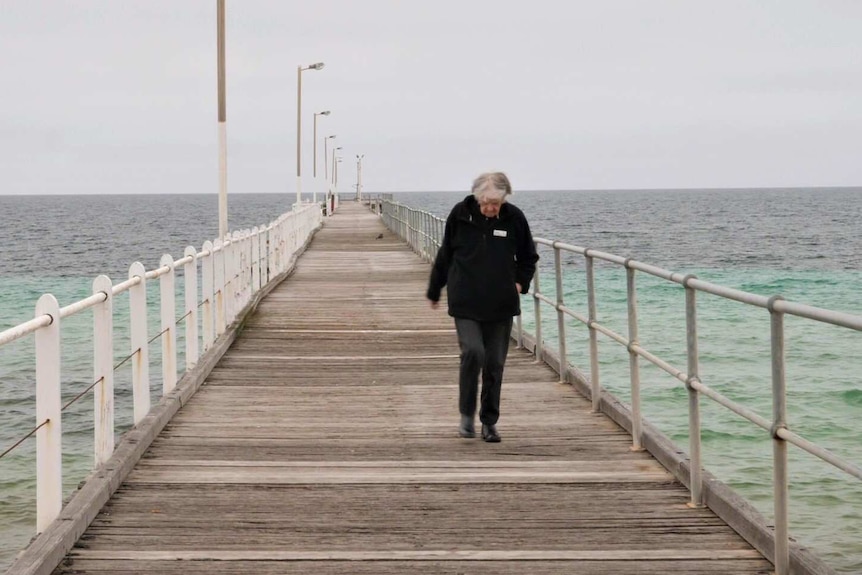 Solitary elderly lady dressed in dark clothing, jacket, walking down jetty with white railing on left, rails on right