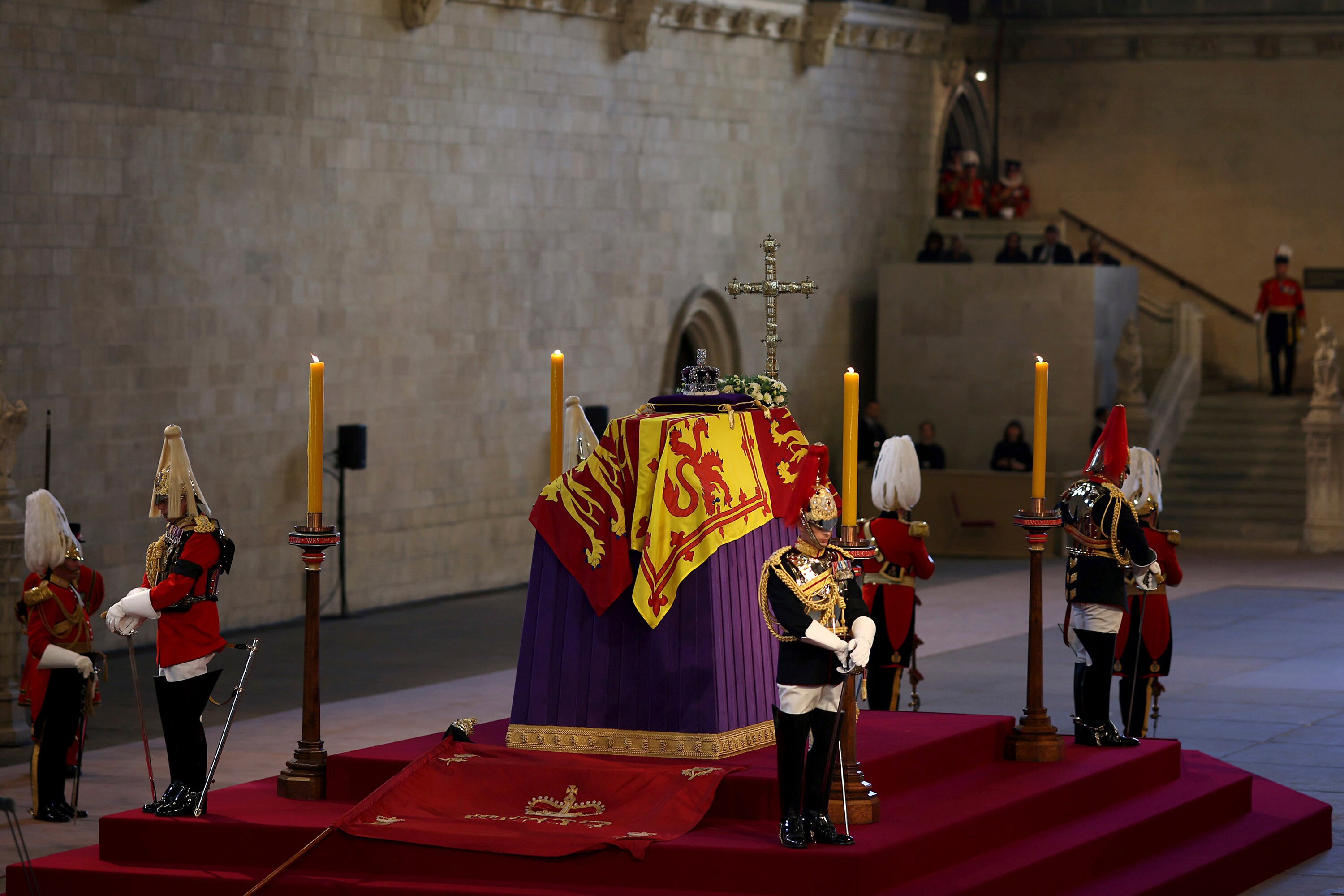A coffin sits on a raised platform surrounded by guards in a hall.