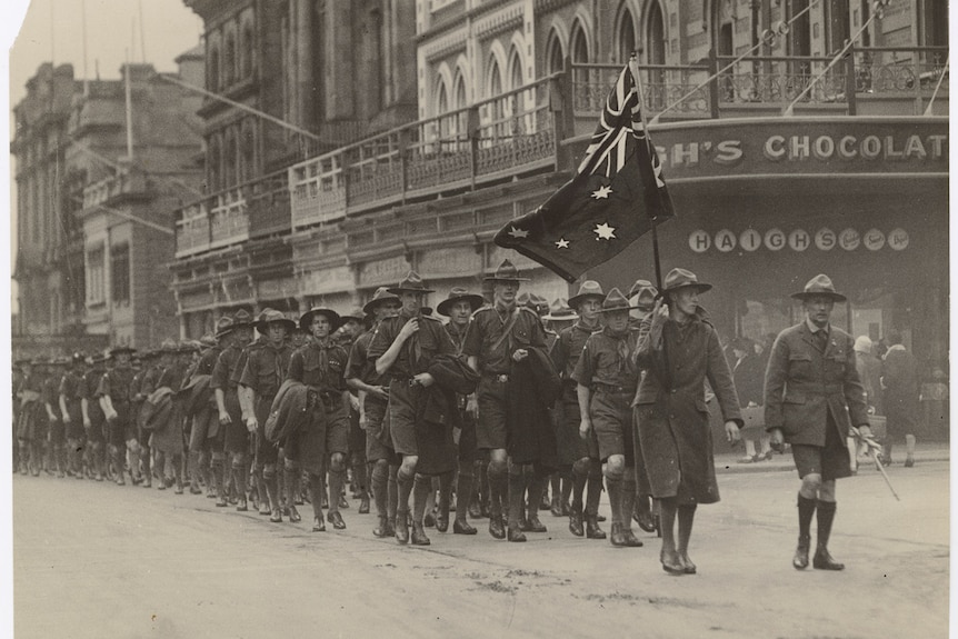 A black and white photo of children wearing scouts uniforms marching through the street, one holding an Australian flag. 