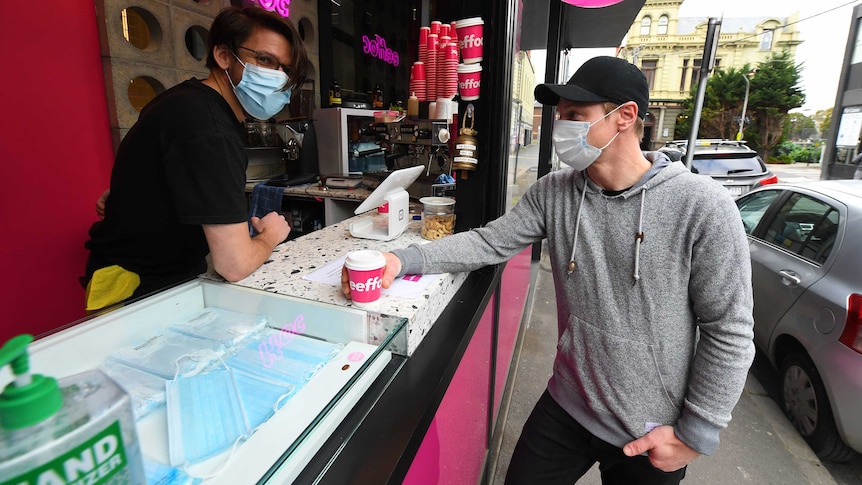 A man stands wearing a face mask in an espresso bar while another man also in a mask takes a coffee.