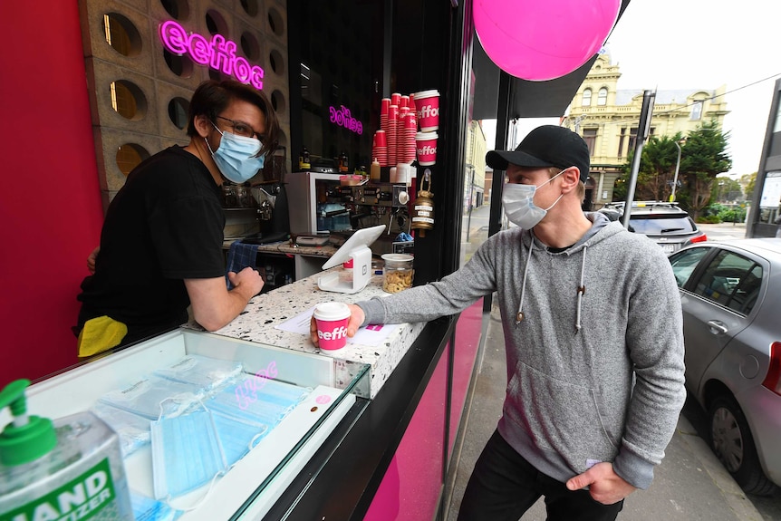 A man stands wearing a face mask in an espresso bar while another man also in a mask takes a coffee.