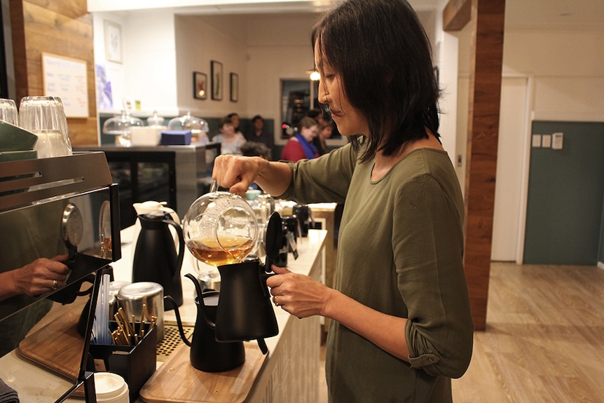 A woman standing at a cafe counter pouring hot tea from a clear glass teapot into a black ceramic teapot.