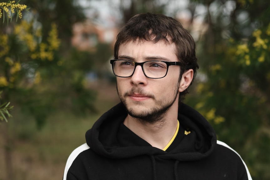 A close up portrait of a teenage boy with glasses, standing with trees and wattle behind him. He is looking to the side.