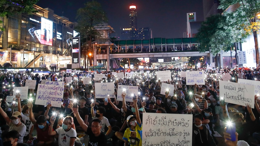 People hold white signs with Thai writing and shine their phone lights surrounded by city builodings. 