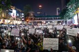 People hold white signs with Thai writing and shine their phone lights surrounded by city builodings. 