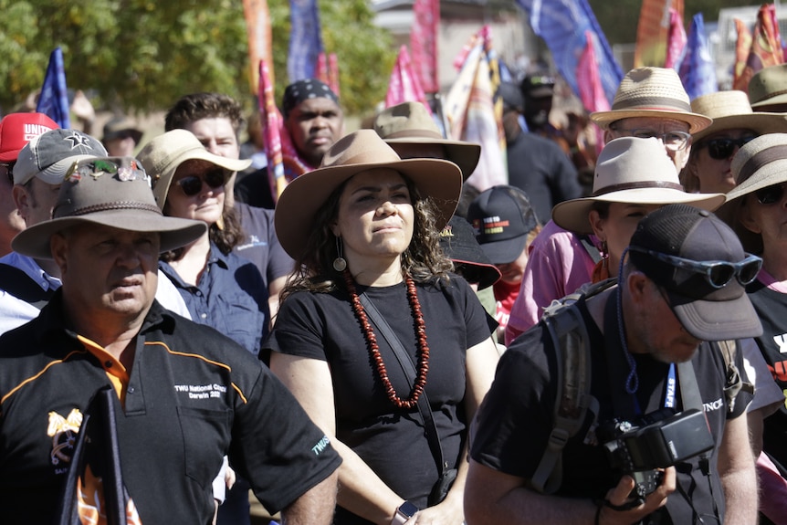 NT Senator Jacinta Price in an Akruba hat and black T-shirt, sitting amid an audience in a bush setting on a sunny day.