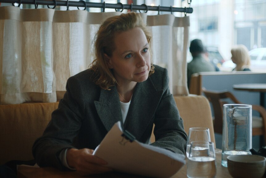 Middle-aged blonde white woman in black business suit sits at diner table holding a wad of paper documents.