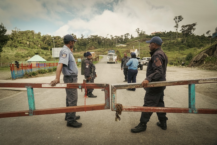 Two police officers in uniform lean against a metal barrier gate across a wide bitumen road