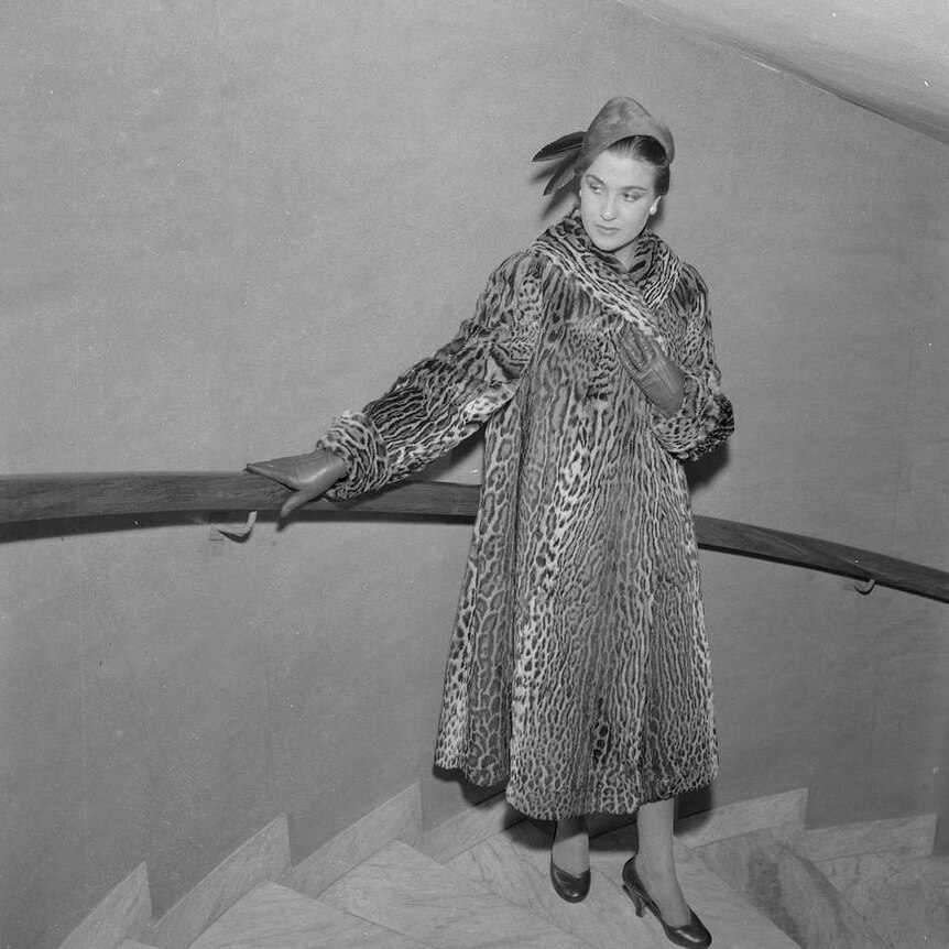 A black and white photo shows a woman in a leopard print coat leans up against a wooden railing and halfway down marble stairs.