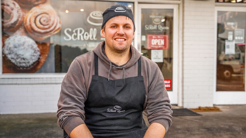 A man in a baker's cap and apron smiles as he sits in front of his scroll shop