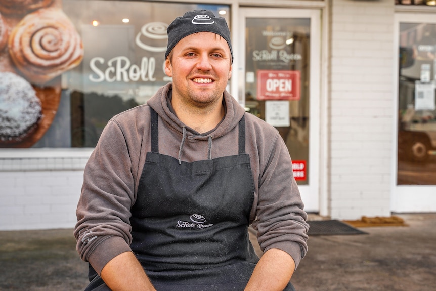A man wearing a chef's hat and apron smiles outside a shop.