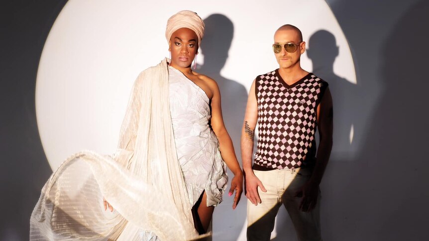 man in white head wrap and white flowing dress and man in sunglasses and checkered vest in front of white cirlce on black wall